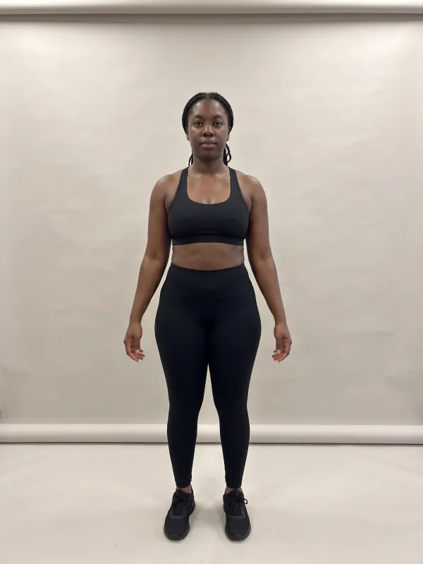 Front-view photo of a woman in a black sports bra and black leggings against a light studio backdrop, with a bottom hourglass body shape: hips wider than the bust and a clearly defined waist.