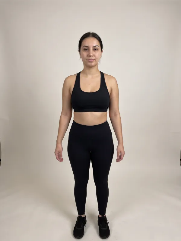Front-view photo of a woman in a black sports bra and black leggings against a light studio backdrop, with an hourglass body shape: bust and hips about the same width and a clearly narrower waist.