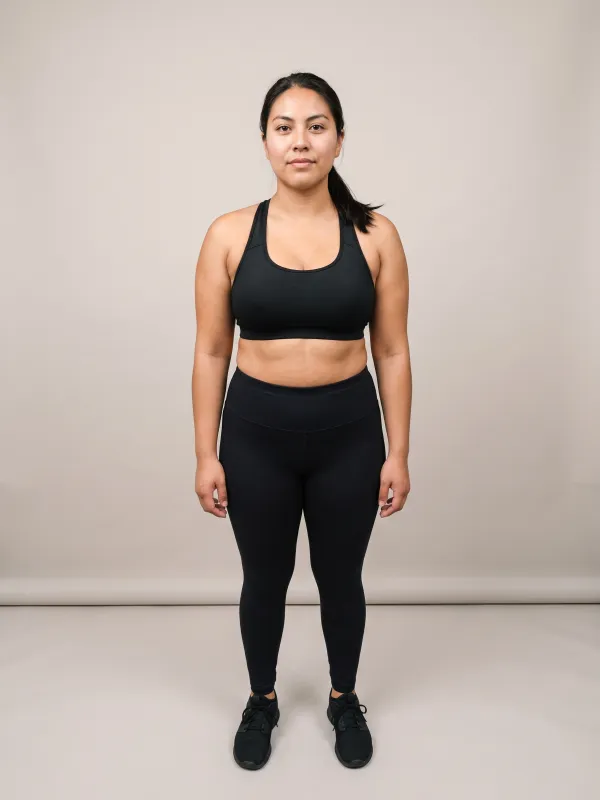 Front-view photo of a woman in a black sports bra and black leggings against a light studio backdrop, with an inverted triangle body shape: broad shoulders and bust tapering to narrower hips.