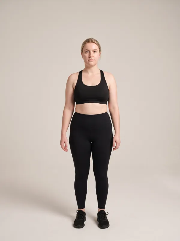 Front-view photo of a woman in a black sports bra and black leggings against a light studio backdrop, with a rectangle body shape: shoulders, bust, waist and hips all similar in width.