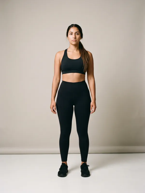 Front-view photo of a woman in a black sports bra and black leggings against a light studio backdrop, with a spoon body shape: hips much wider than the bust, flaring out sharply just below a defined waist.