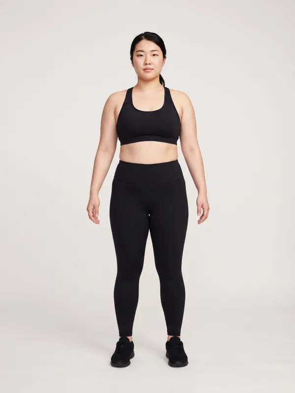 Front-view photo of a woman in a black sports bra and black leggings against a light studio backdrop, with a top hourglass body shape: bust and shoulders wider than the hips and a clearly defined waist.