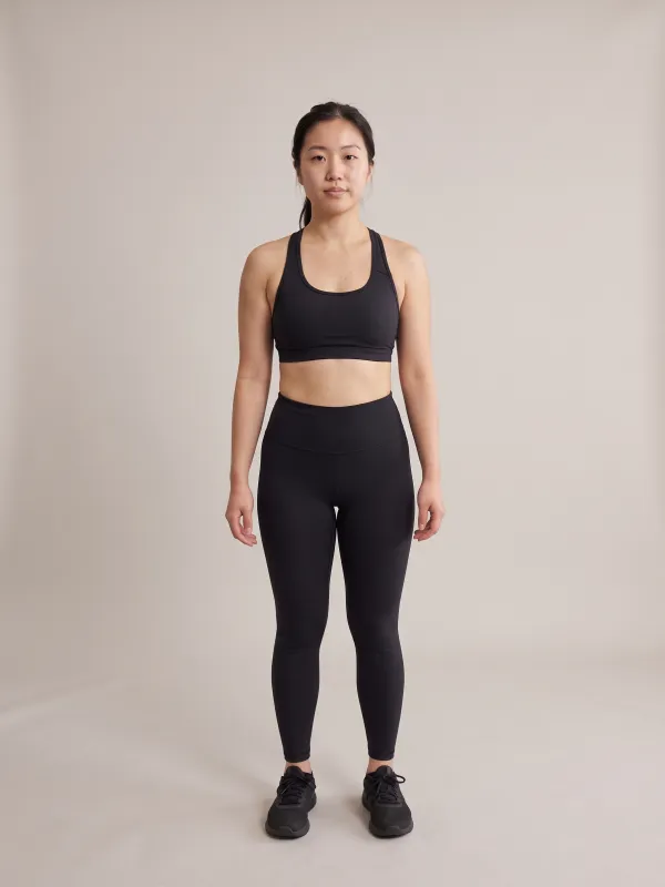 Front-view photo of a woman in a black sports bra and black leggings against a light studio backdrop, with a triangle body shape: hips and thighs wider than the bust, with only a little waist definition.
